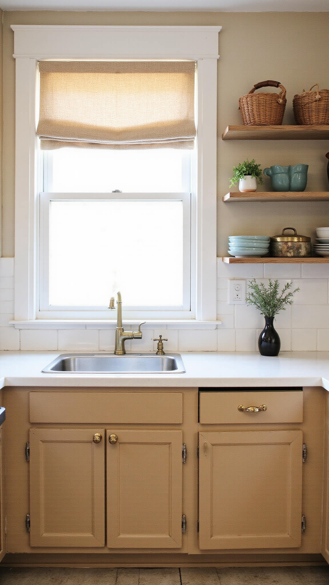 Warm beige cabinets in a compact 12x14ft kitchen with brass hardware, open shelving, vintage decor, and rattan baskets, lit by afternoon sun through Roman shades.