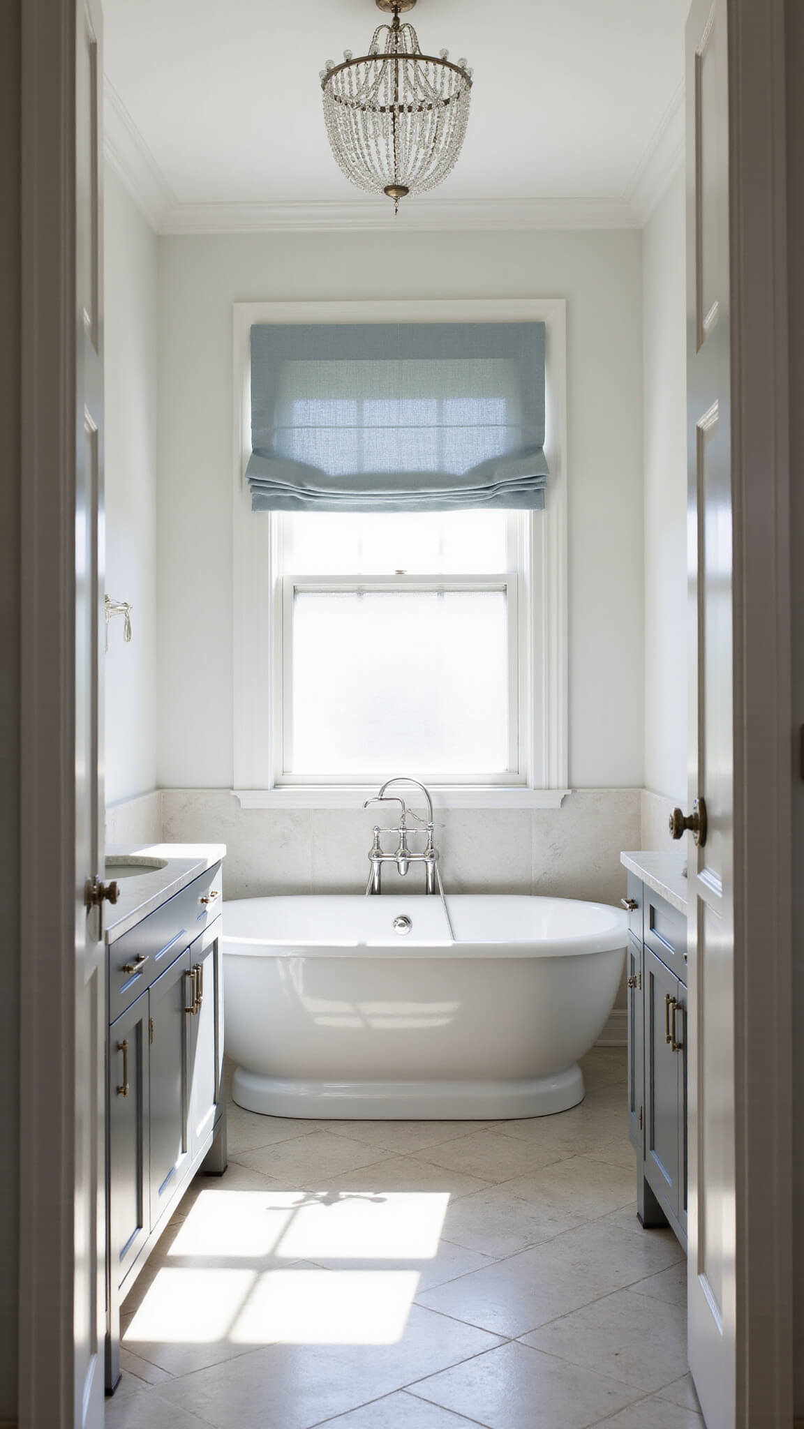 Wide-angle view of a luxurious transitional bathroom with a white freestanding tub on marble herringbone floors, double gray vanity with Carrara marble top, and morning light through frosted window.