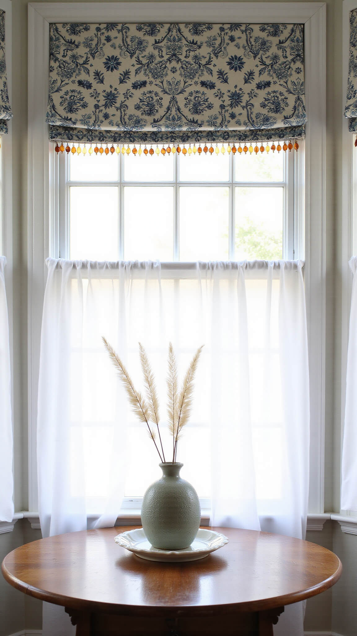 Cozy 8x8ft breakfast nook with bay windows, layered window treatments, a distressed wood table with pampas grass in a ceramic vase, bathed in soft early morning light.