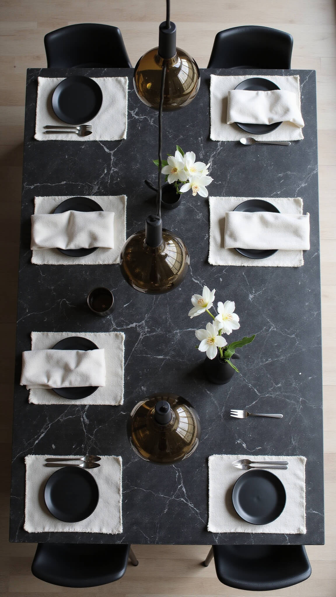 Overhead view of a black marble kitchen island with pendant light reflections, black bar stools, white placemats, matte black dinnerware, and white orchids in morning light.