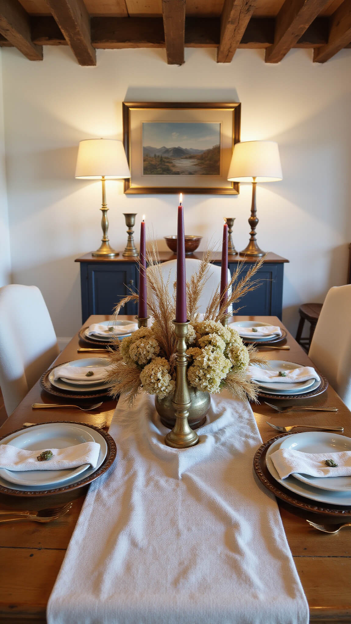 Twilight dining room tablescape with rustic harvest table, dried floral centerpiece in copper vase, brass candlesticks, and pottery chargers under wooden beams.