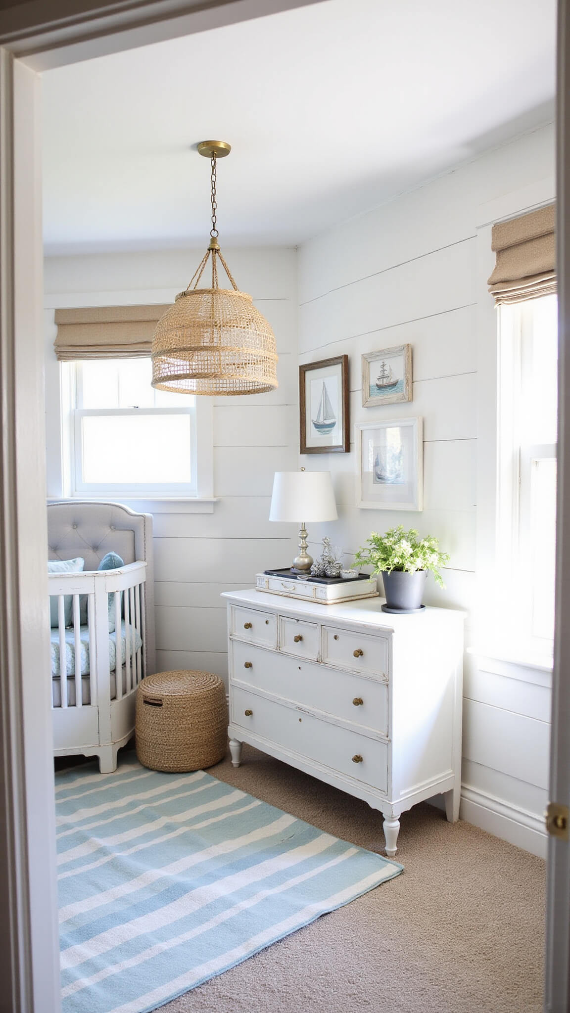 Coastal-themed 12x14 nursery with white shiplap wall, light grey crib, rope pendant light, weathered white dresser, striped rug, natural linen shades, vintage boat prints, jute pouf, and seagrass baskets in bright midday lighting.