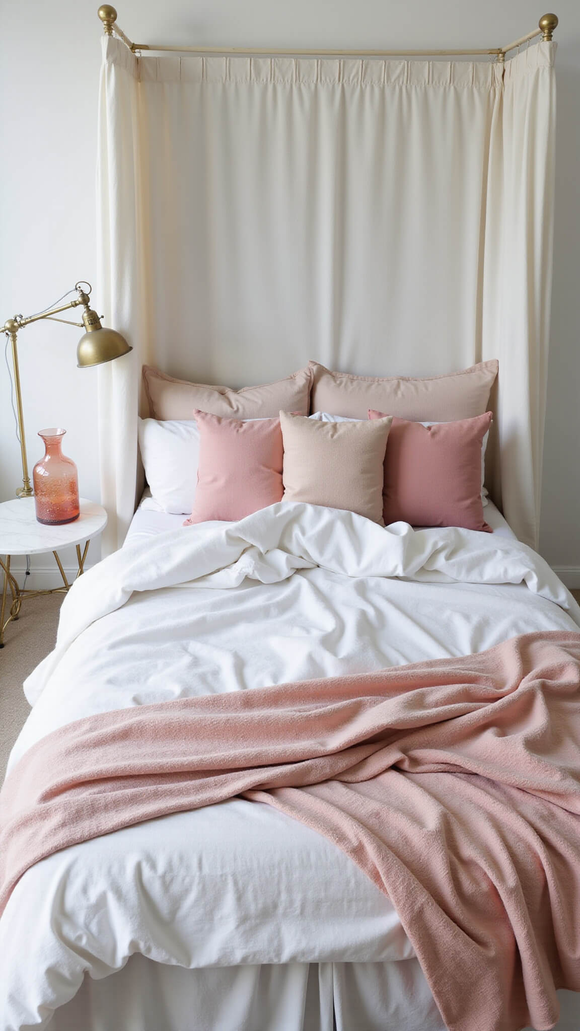 Overhead view of a white canopy bed with rumpled linen bedding, blush pink throw, assorted pink pillows, brass reading lamp, and marble side table with vintage pink carafe.