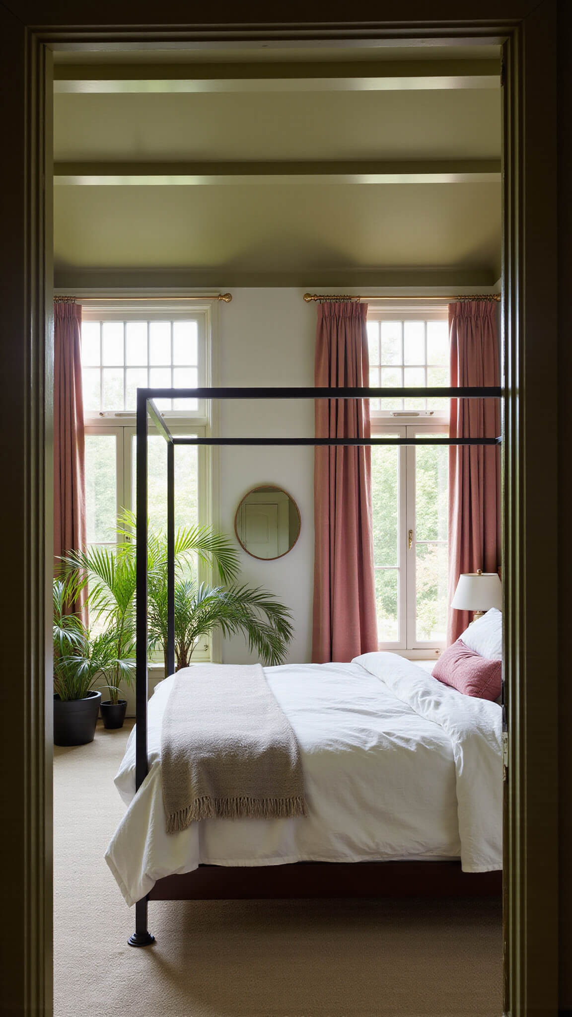 Late afternoon light fills a cozy bedroom with olive green ceiling beams, pink velvet curtains, and a modern four-poster bed with rumpled linen bedding, viewed through a doorway.