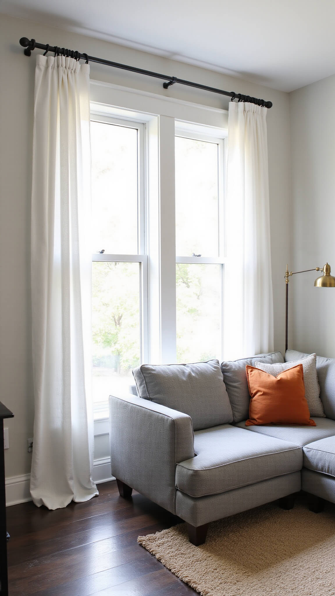 Bright, airy living room with high ceilings, grey sectional, sheer white curtains, and sunlight streaming onto dark hardwood floors.