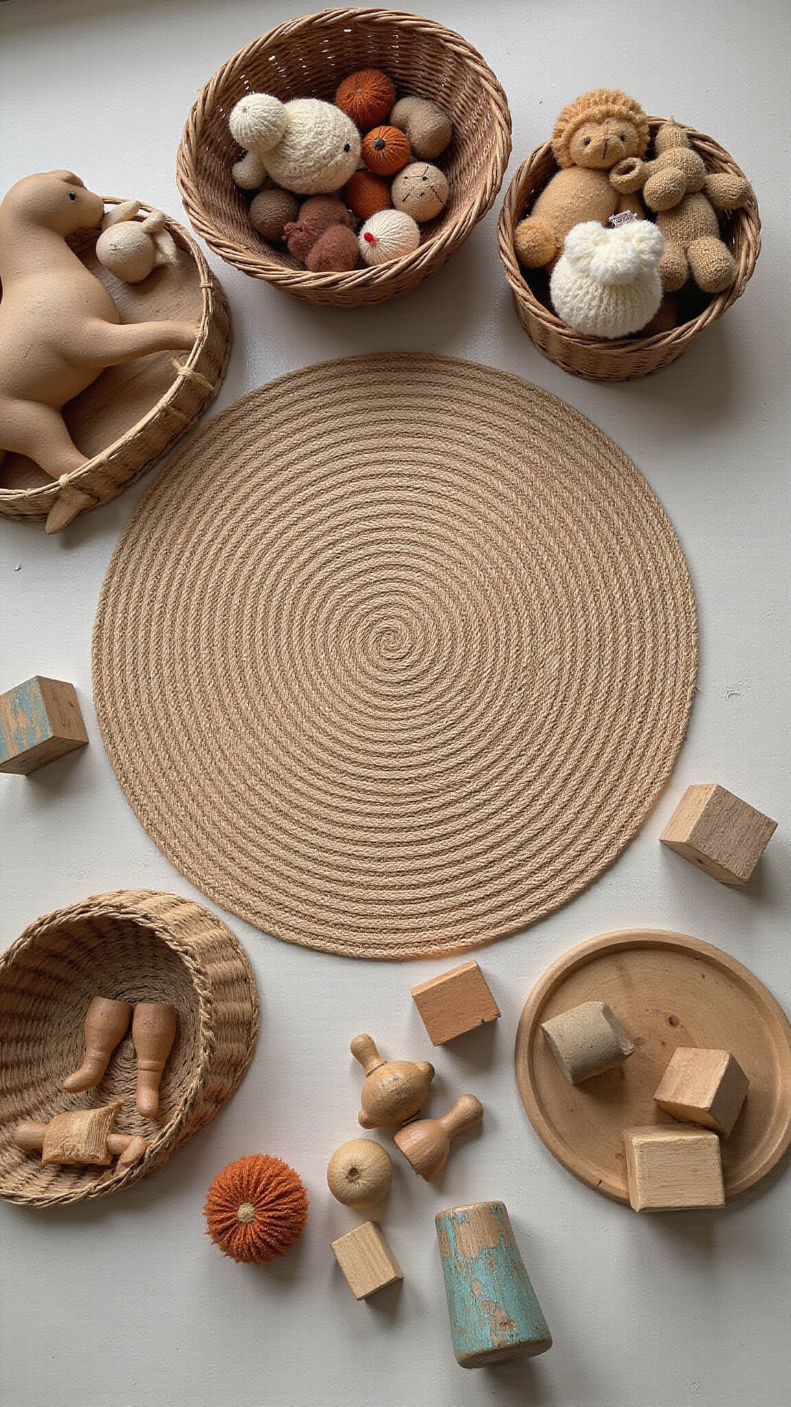 Overhead view of a vintage nursery play area with a round braided wool rug, wooden rocking horse, faded building blocks, and wicker baskets filled with handmade dolls and knit animals, all lit by soft natural light.
