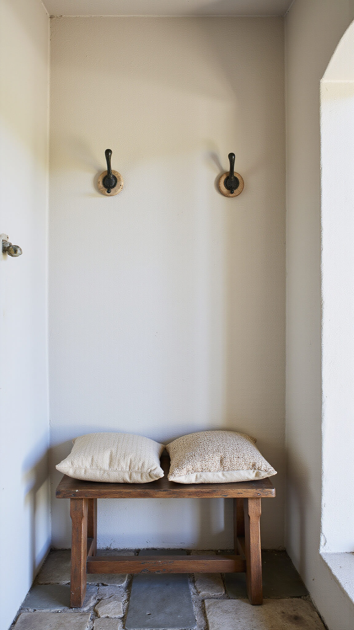 Wabi Sabi entryway with textured plaster walls, antique wooden bench, handwoven cushions, ceramic coat hooks, and worn stone flooring in muted natural tones.