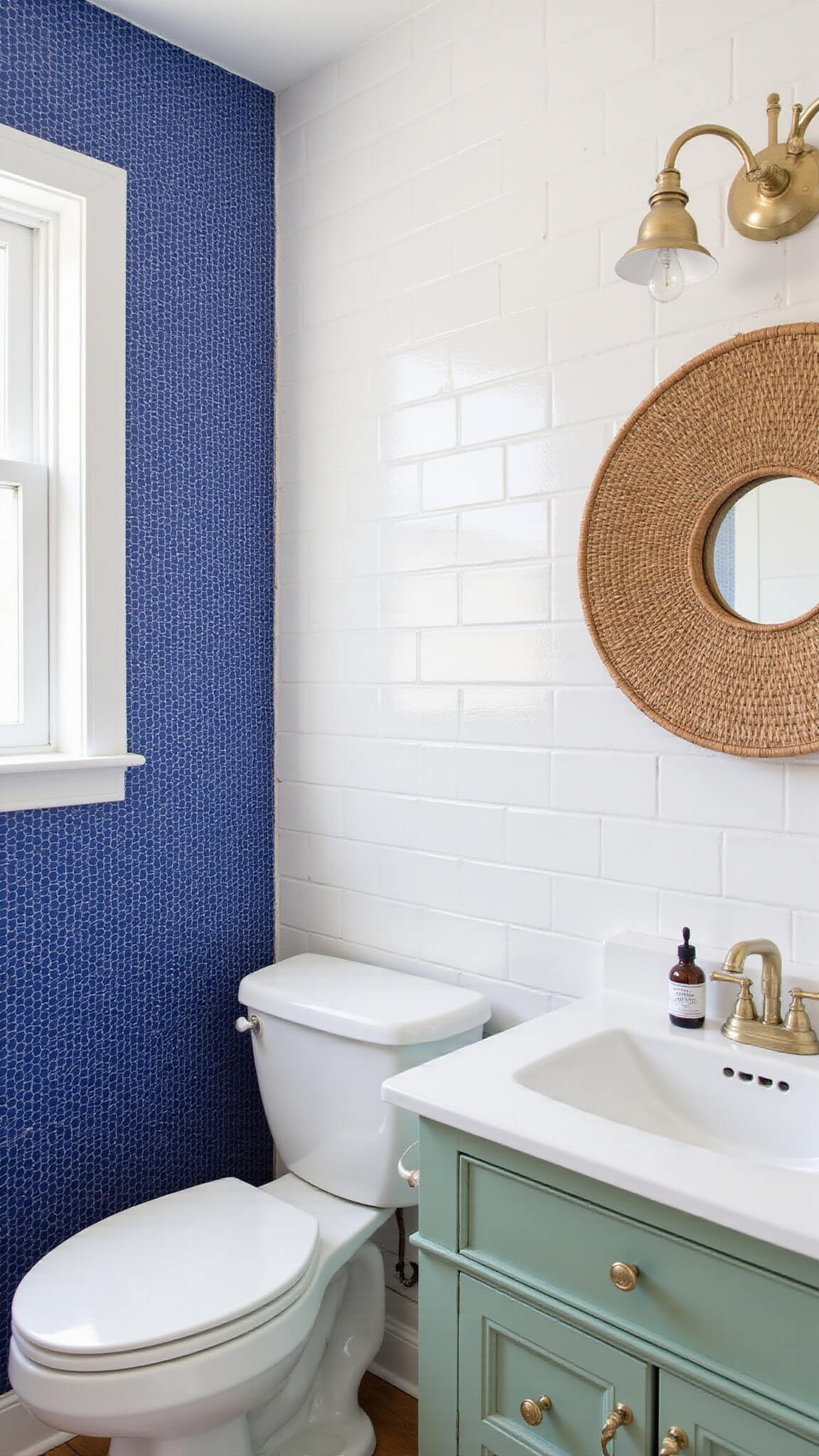Small bathroom with white subway tiles and cobalt blue penny tile accent wall, featuring pedestal sink, round rattan mirror, sage green DIY-painted vanity with brass hardware, and vintage accessories.