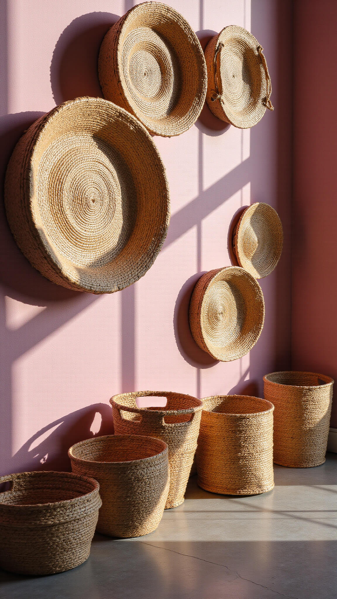 Low-angle view of a 12ft wall displaying vintage woven baskets in natural tones on soft pink wallpaper, with late afternoon light casting geometric shadows.
