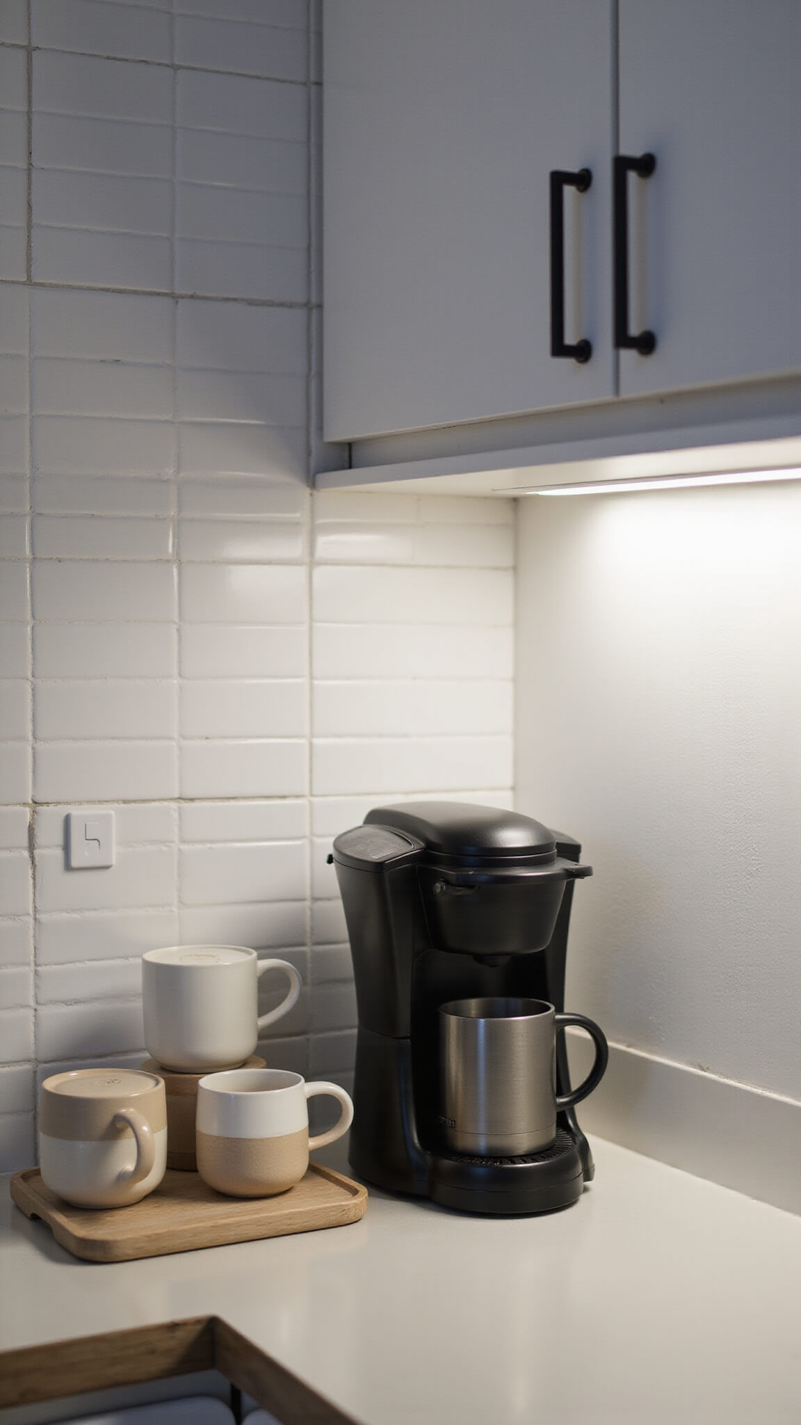Nordic kitchen coffee station at dusk with matte black coffee maker, ceramic mugs on wooden tray, and warm under-cabinet lighting.