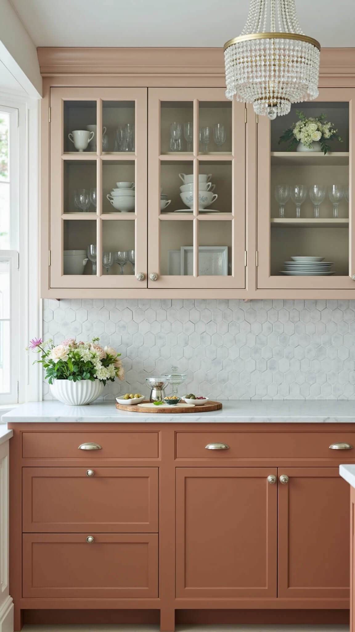 High-angle view of a 13x15ft transitional kitchen with terracotta lower cabinets, glass-front uppers, marble hexagon backsplash, crystal chandelier, vintage glassware, and fresh flowers at midday.