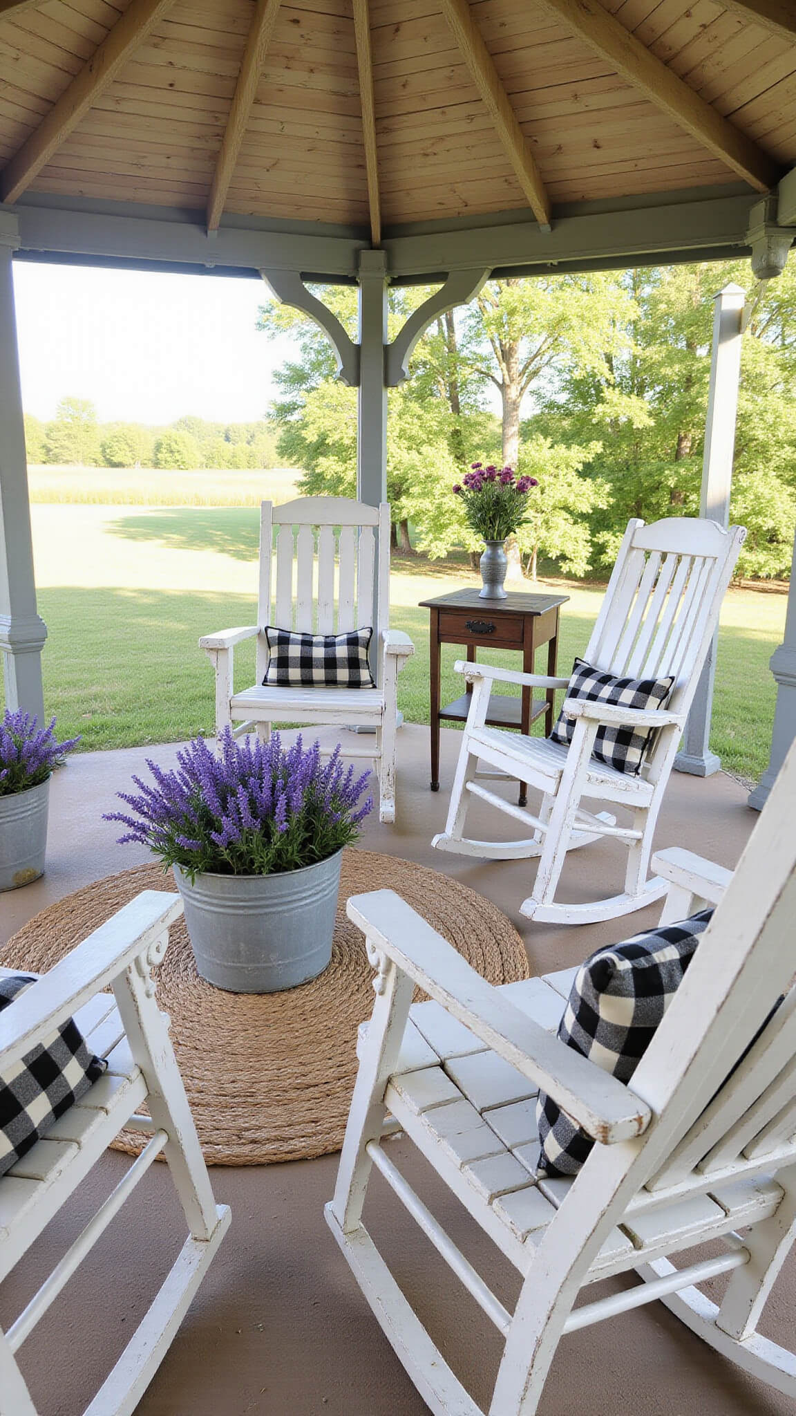 Farmhouse-style gazebo with distressed white furniture, oversized rockers, vintage tea cart, and metal planters of lavender and roses; black and cream cushions add charm under rustic ceiling beams in late afternoon light.
