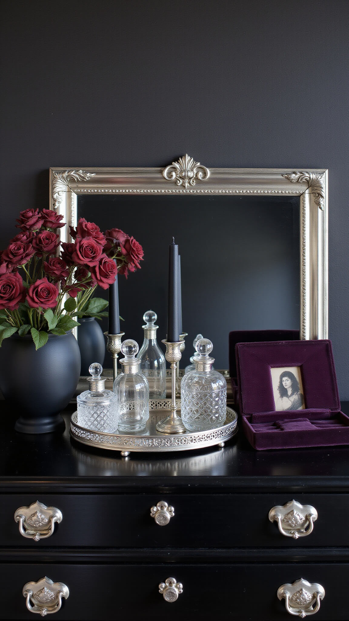 Close-up of antique black gothic dresser with silver hardware, vintage mirror tray holding crystal perfume bottles, black candles in silver holders, dried roses in matte black vase, ornate silver frame with vintage photo, and dark purple velvet jewelry box against textured black wallpaper in soft morning light.