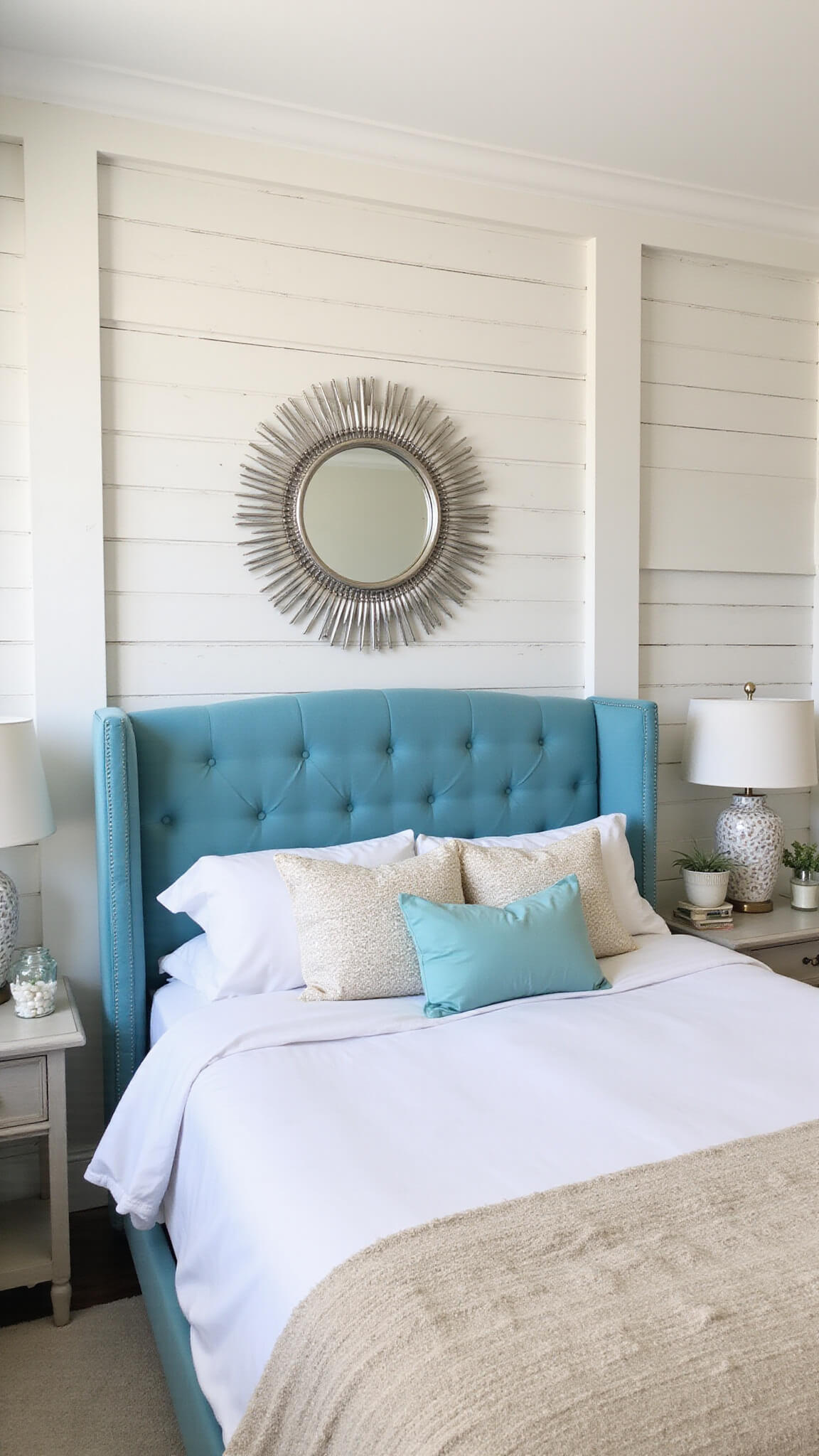 Coastal glam bedroom with sea glass blue headboard, white shiplap accent wall, textured bedding in beige and aqua, silver sunburst mirror, and driftwood nightstands under soft mid-morning light.