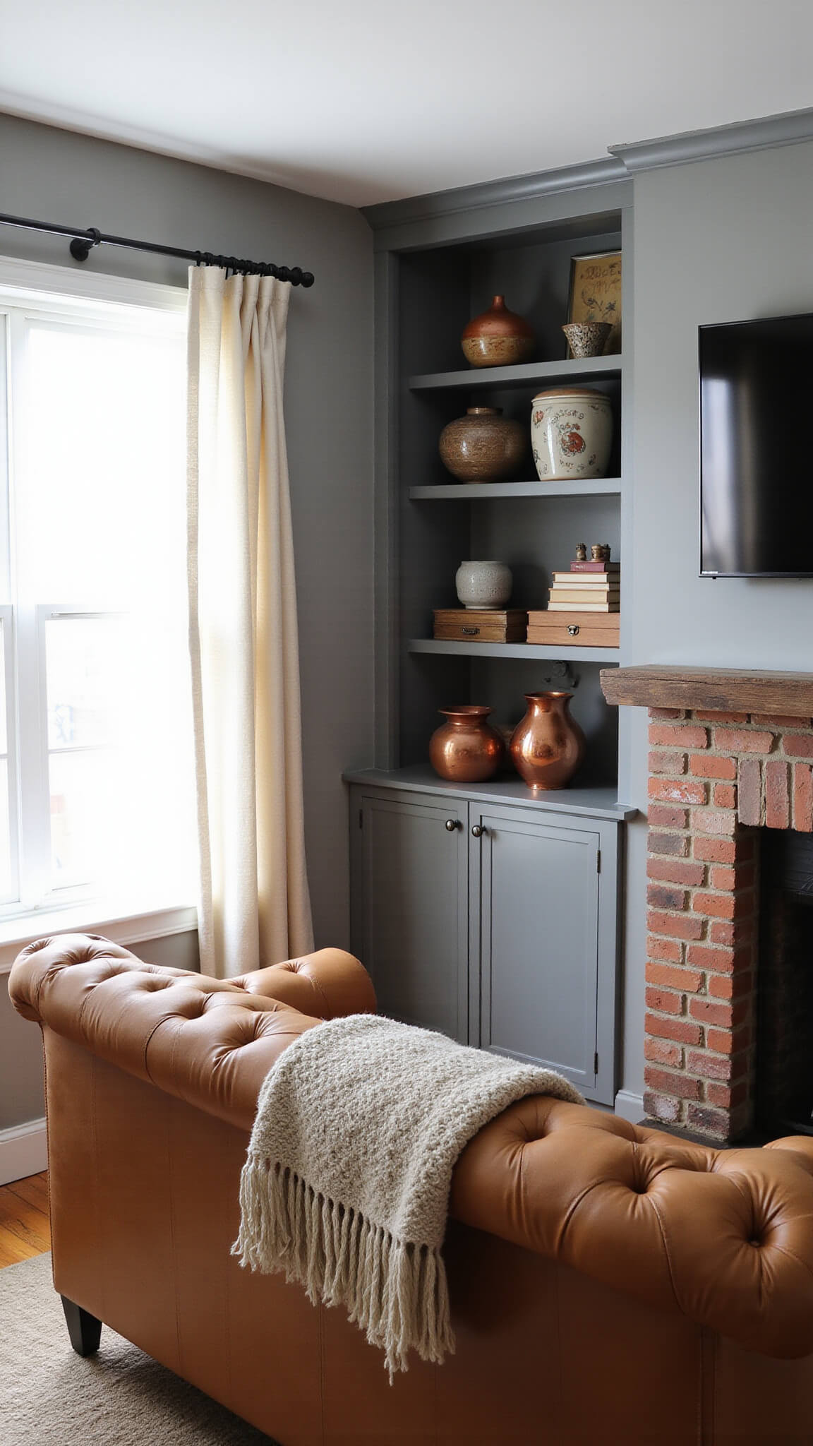 Cozy morning scene in a small living space with a camel leather Chesterfield sofa, brick fireplace, wooden mantel, built-in bookshelves, and soft light filtering through cafe curtains.