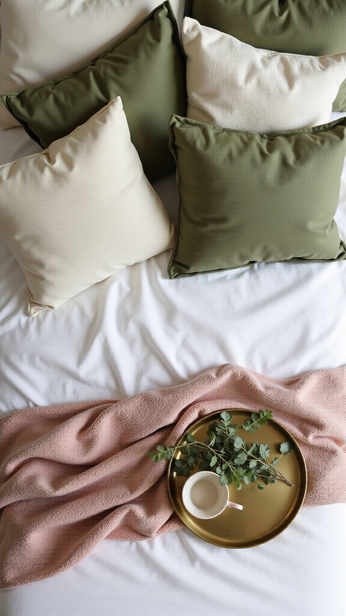 Overhead view of olive green linen bedding with sage, moss, and cream pillows, dusty pink throw, and brass tray holding ceramic mug and eucalyptus.