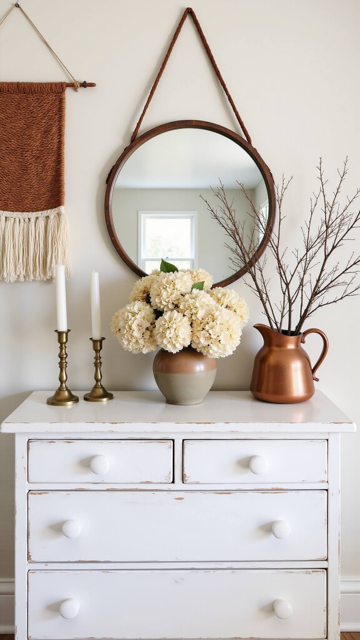 Modern farmhouse bedroom vignette with white distressed dresser, circular mirror, brass candlesticks, dried hydrangeas in earthenware vase, rust and cream woven wall hanging, and vintage copper pitcher with preserved branches.