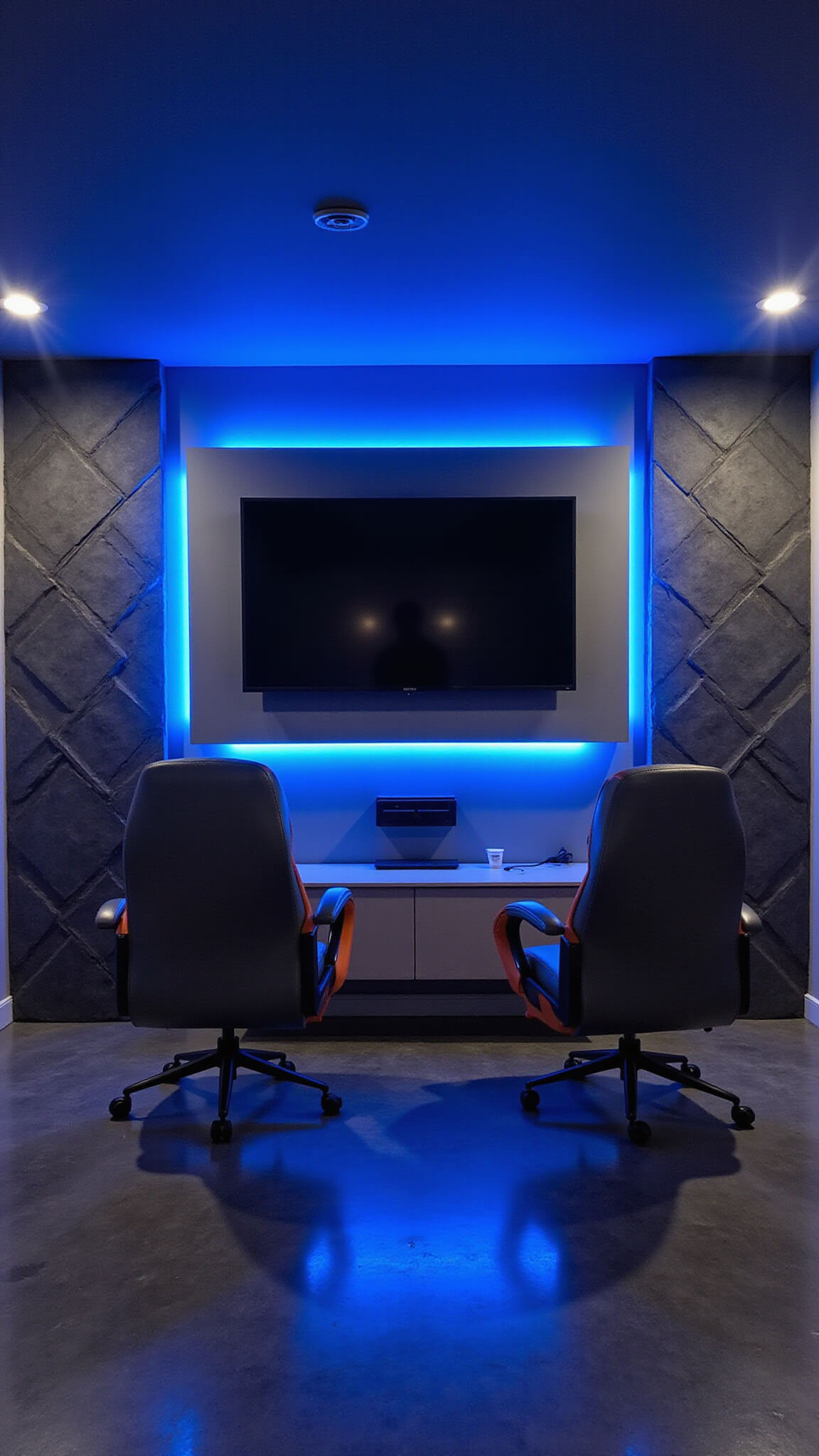 Ground-level view of a modern gaming room at night with electric blue LED lighting, geometric charcoal acoustic panels, gunmetal gray gaming chairs with neon orange accents, and polished concrete floors reflecting ambient glow.