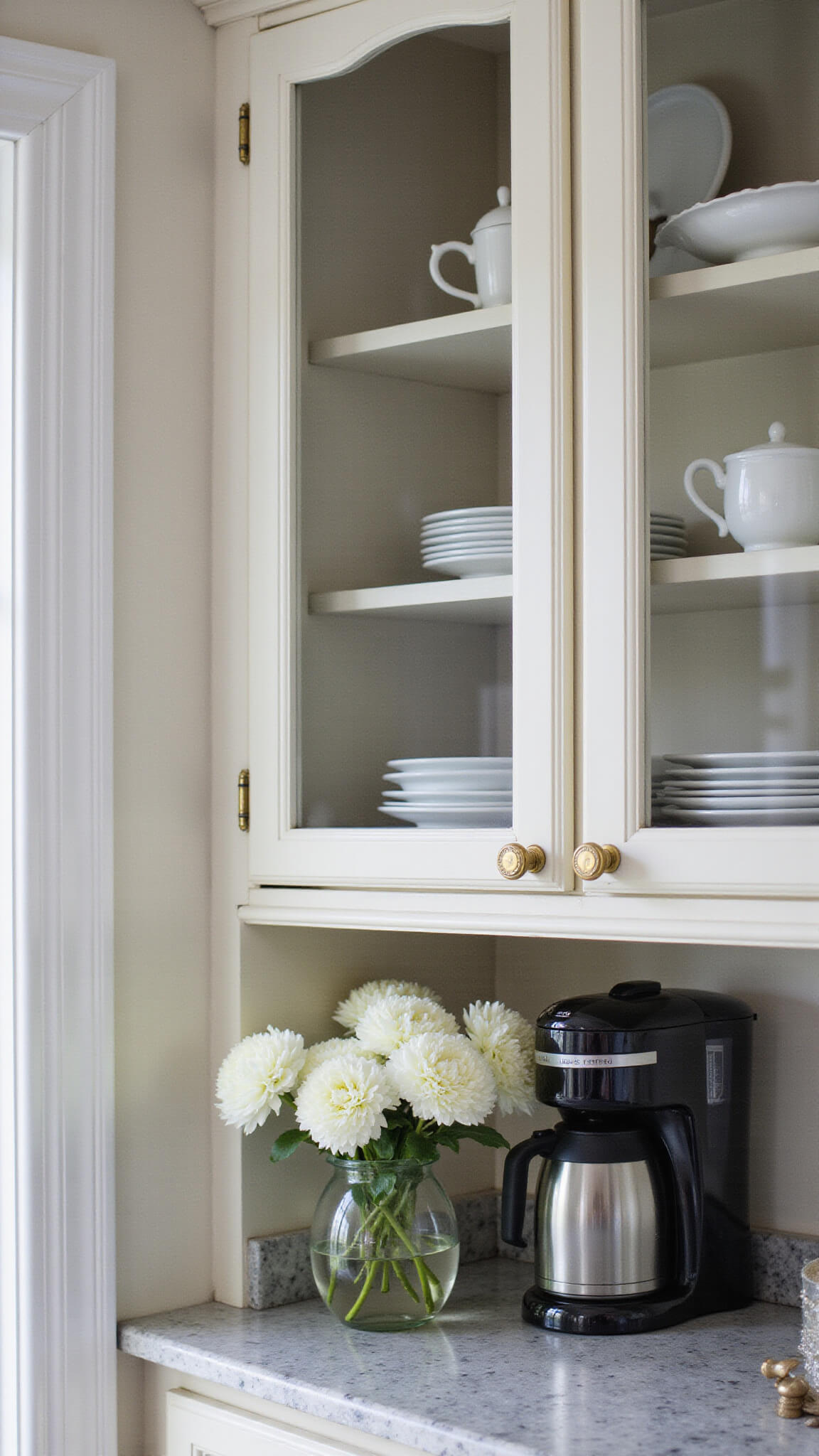 Swiss Coffee cabinet with glass doors and brass hardware in morning light, displaying white pottery, marble countertop, coffee station, and white blooms in clear vase.