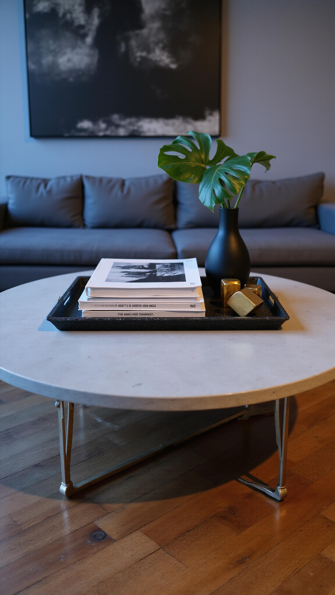 Contemporary urban loft interior with concrete coffee table on walnut flooring, styled with black shagreen tray, monochrome art books, matte black vase with monstera leaf, and brass objects, lit by cool natural and warm LED light.