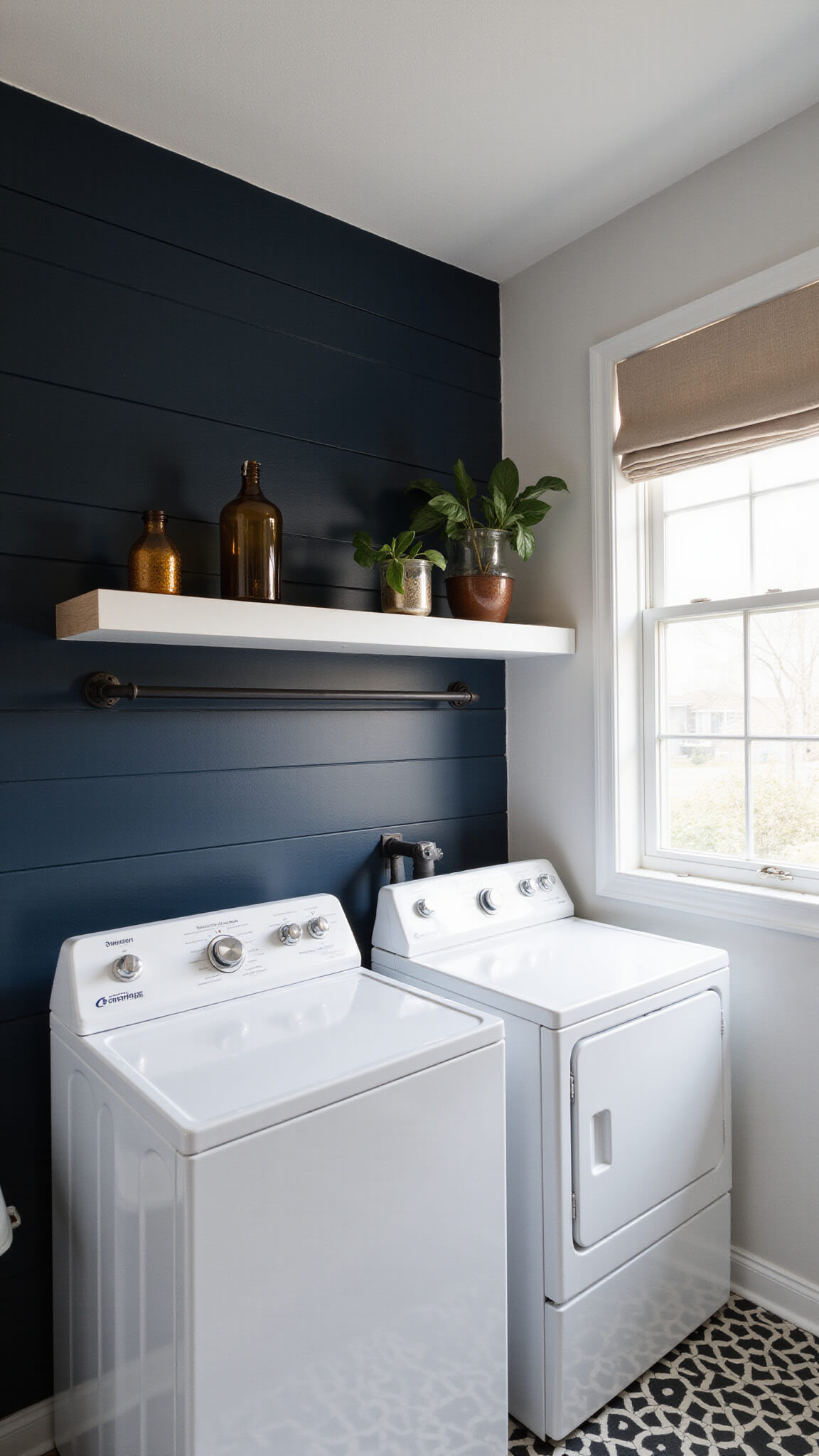 Modern minimalist laundry room with stacked white appliances against navy shiplap wall, floating oak shelves, amber glass decor, monstera plant, matte black hardware, geometric floor tiles, and soft mid-morning light.
