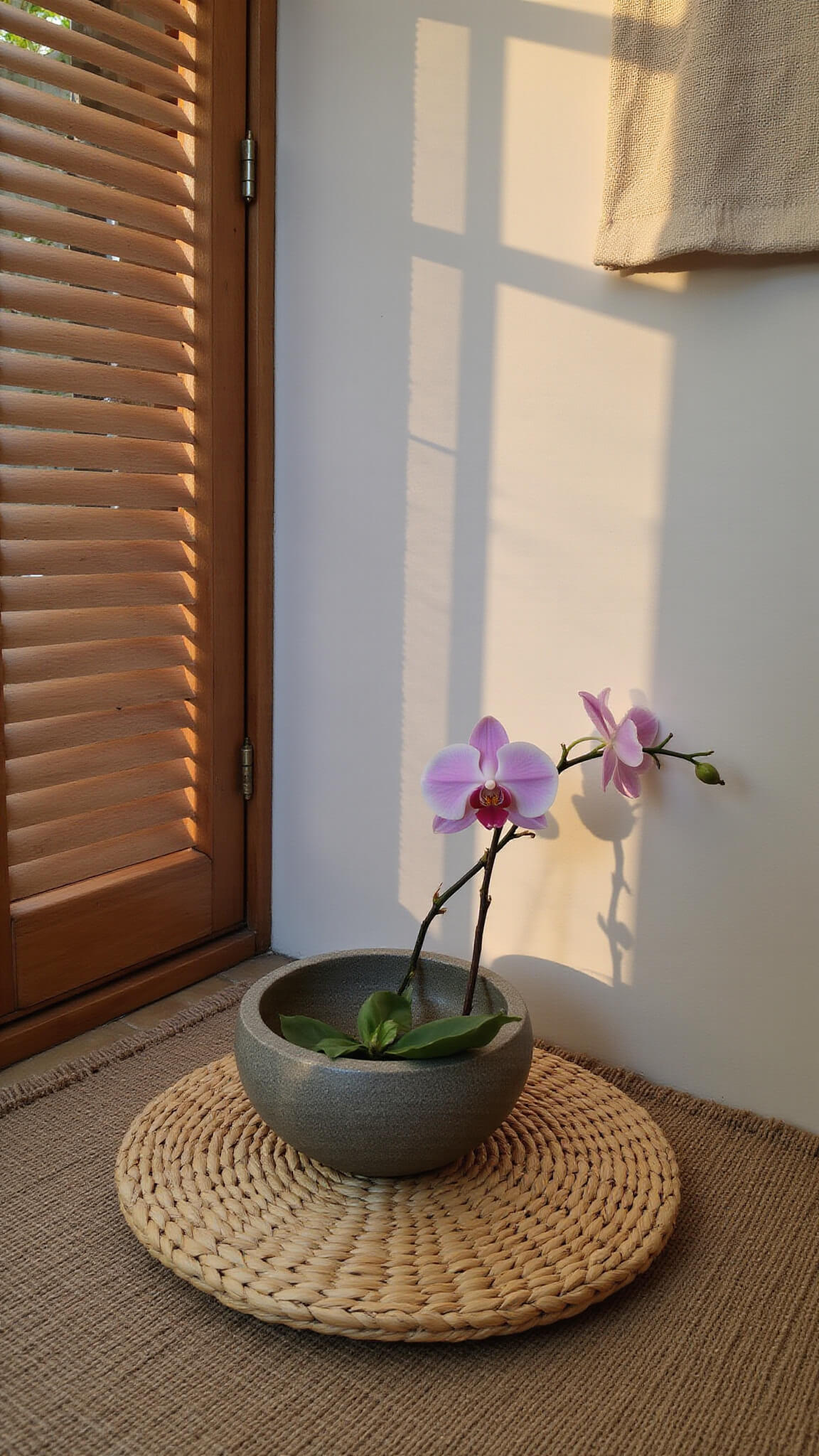 Meditation corner with rattan cushion on bamboo mat, orchid in stone bowl, and linen wall hanging, lit by dusk light through wooden screen.