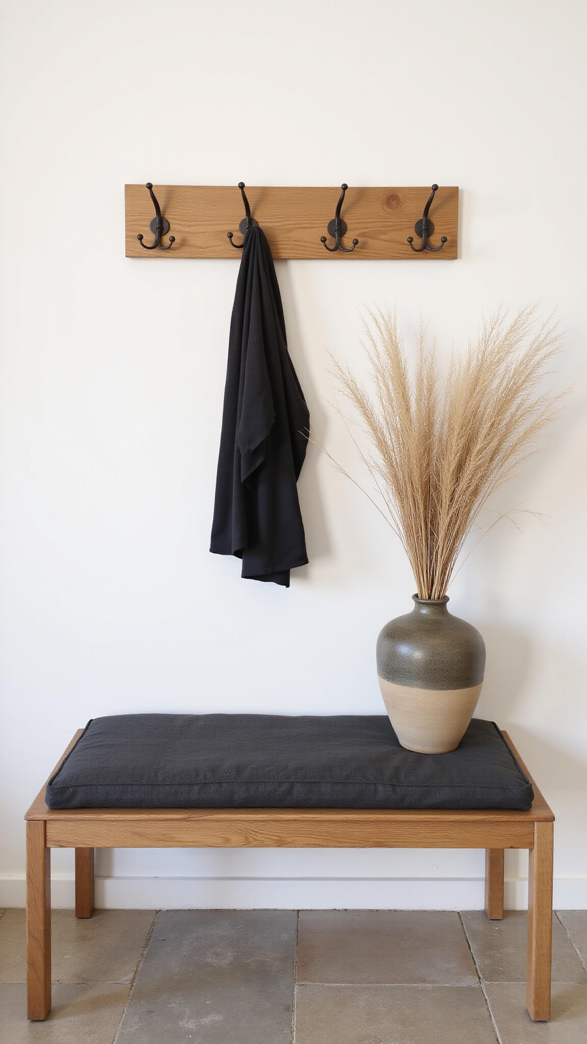 Entryway with floating oak bench, charcoal cushion, metal hooks, pampas grass in ceramic vessel, and natural stone flooring against white walls.
