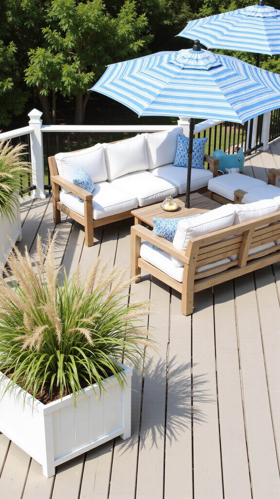 Sunlit coastal deck with teak sectional, white cushions, striped umbrellas, and swaying grasses overlooking ocean tones.