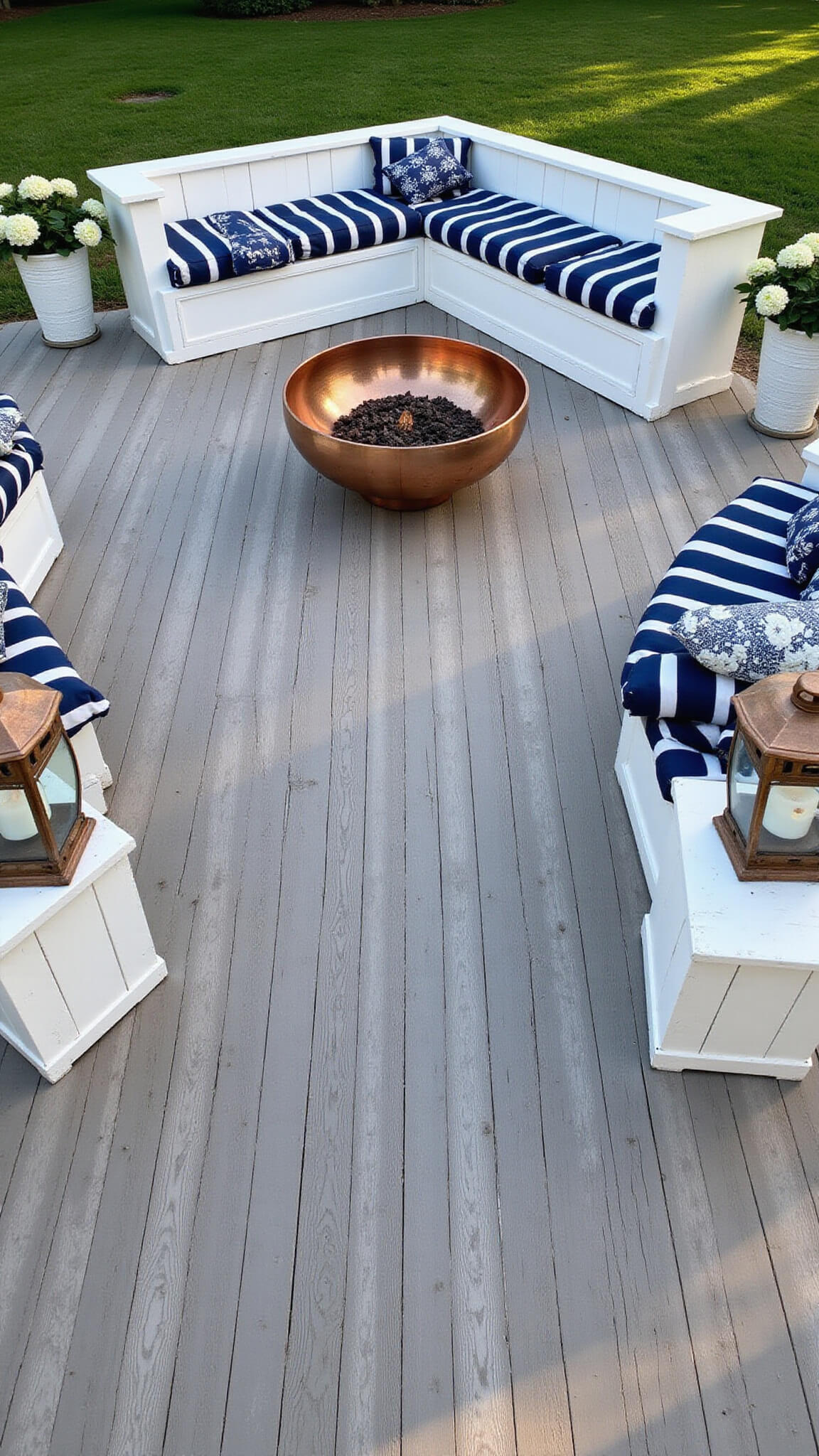 Bird's-eye view of a coastal 18x18ft raised deck with white-washed benches around a copper fire bowl, navy and white cushions, nautical decor, and potted hydrangeas in late afternoon light.