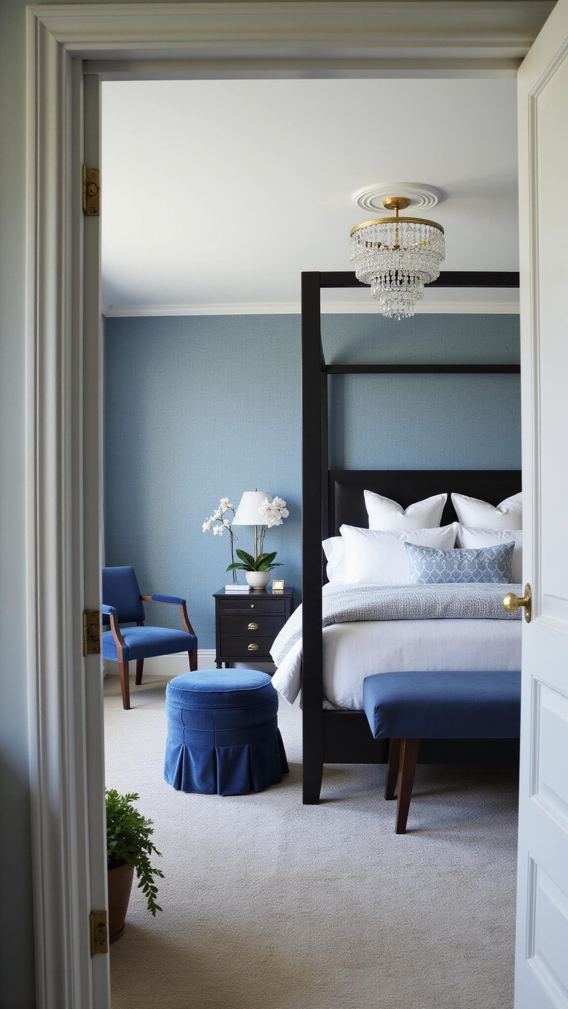 Luxury 15x17ft bedroom with tall ceilings, blue grasscloth wallpaper, black four-poster bed with white and blue linens, blue velvet chairs, crystal chandelier, and orchids, viewed from doorway with tilt-shift effect.