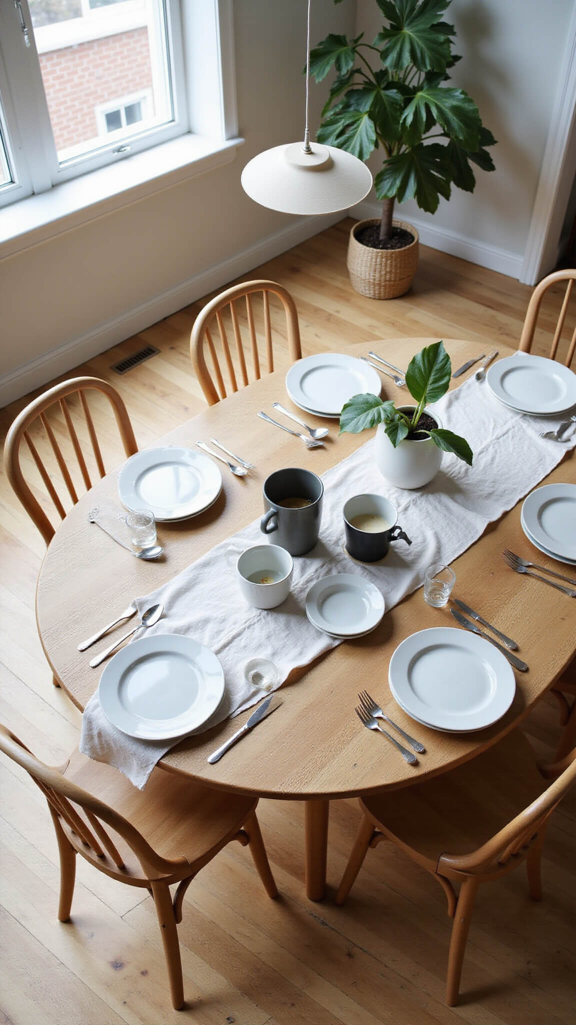 Overhead view of round oak dining table with wishbone chairs, linen tablecloth, white and gray ceramics, paper pendant light, and fiddle leaf fig in natural light.