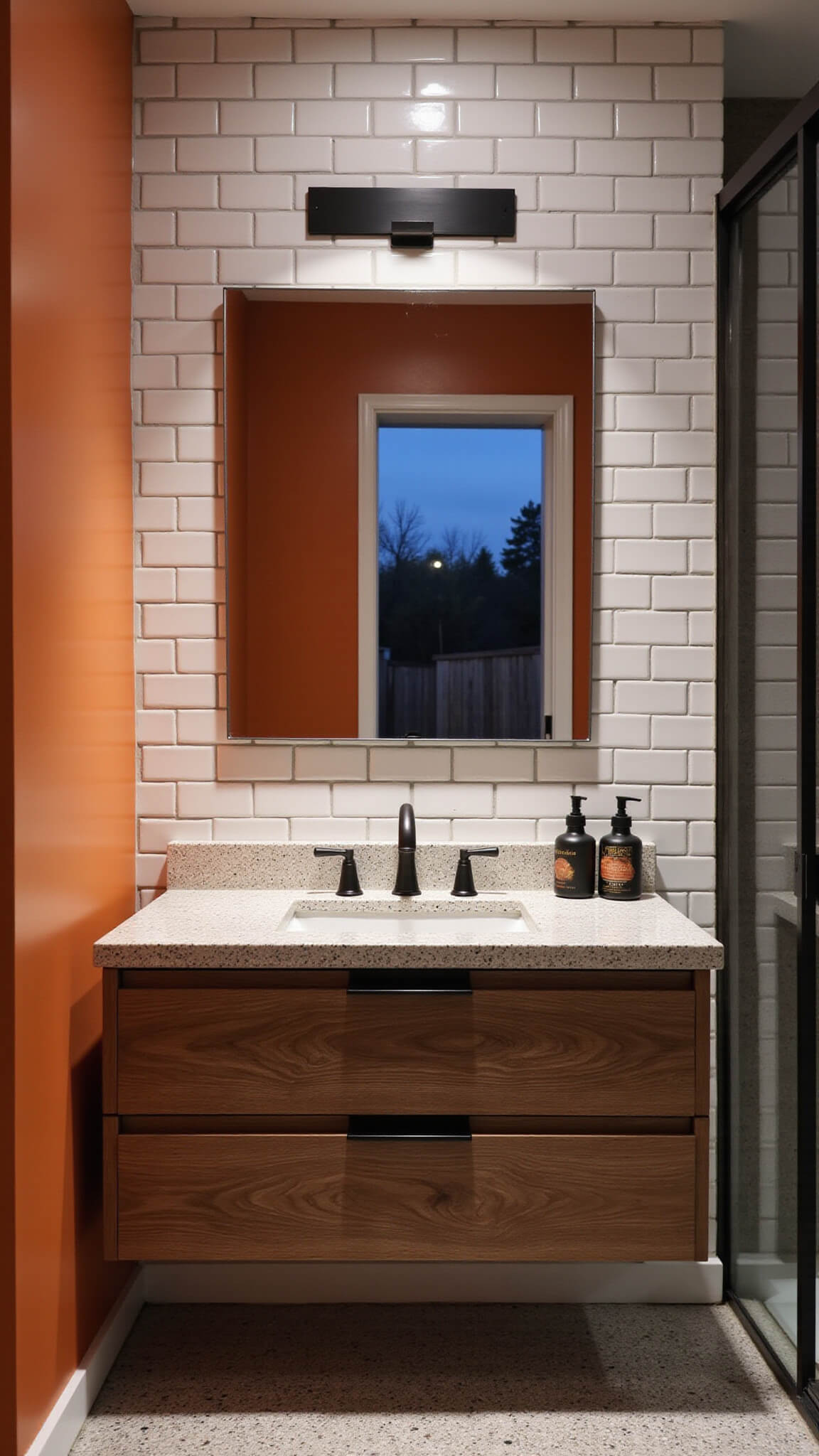 Twilight bathroom with burnt orange accent wall, white subway tiles, walnut floating vanity, matte black hardware, terrazzo flooring, and moody lighting.