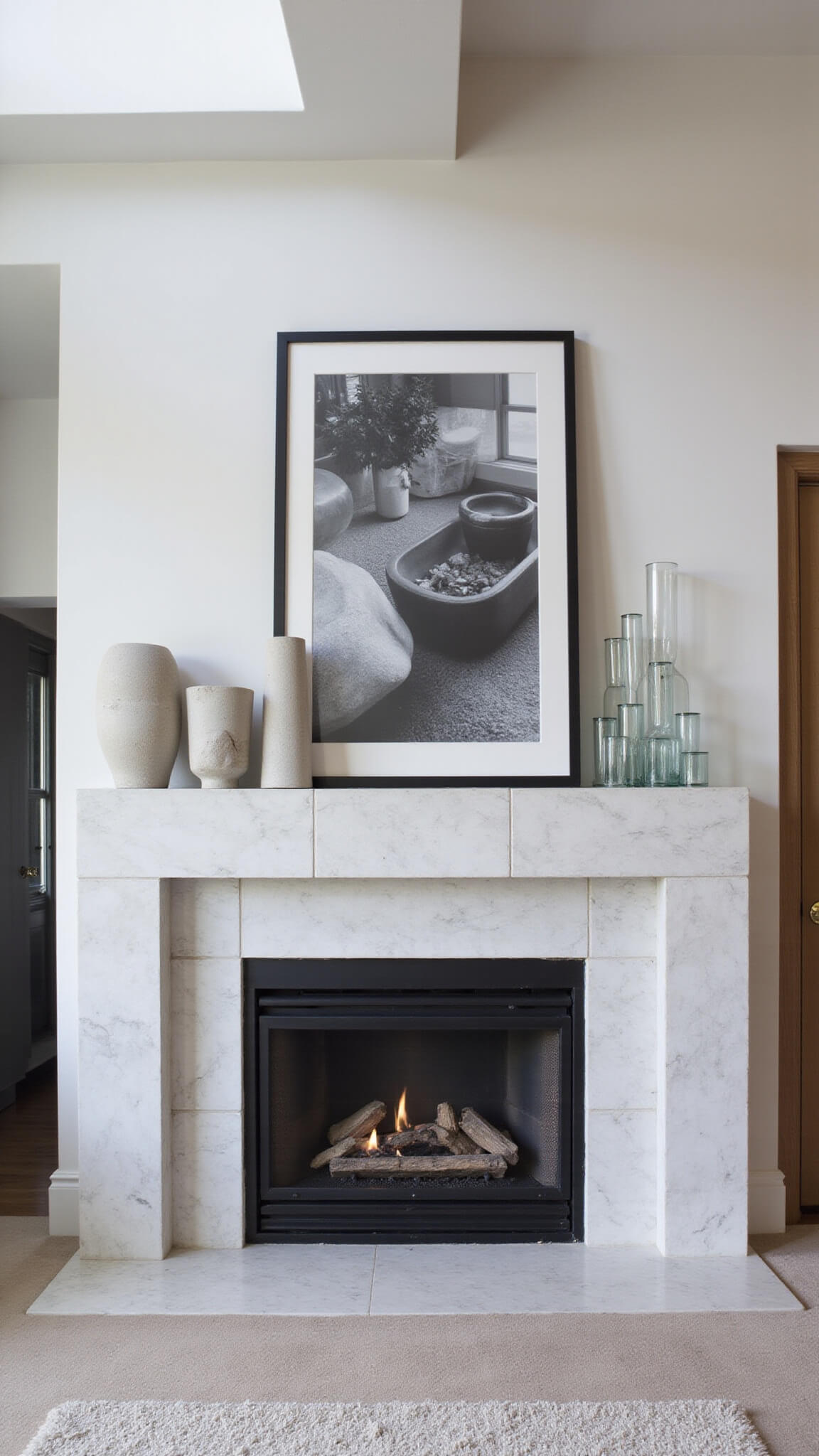 High-angle view of modern open-concept great room with floor-to-ceiling stone fireplace, white marble mantle displaying concrete sculptures, black and white photography, and glass cylinders, lit by natural skylight.