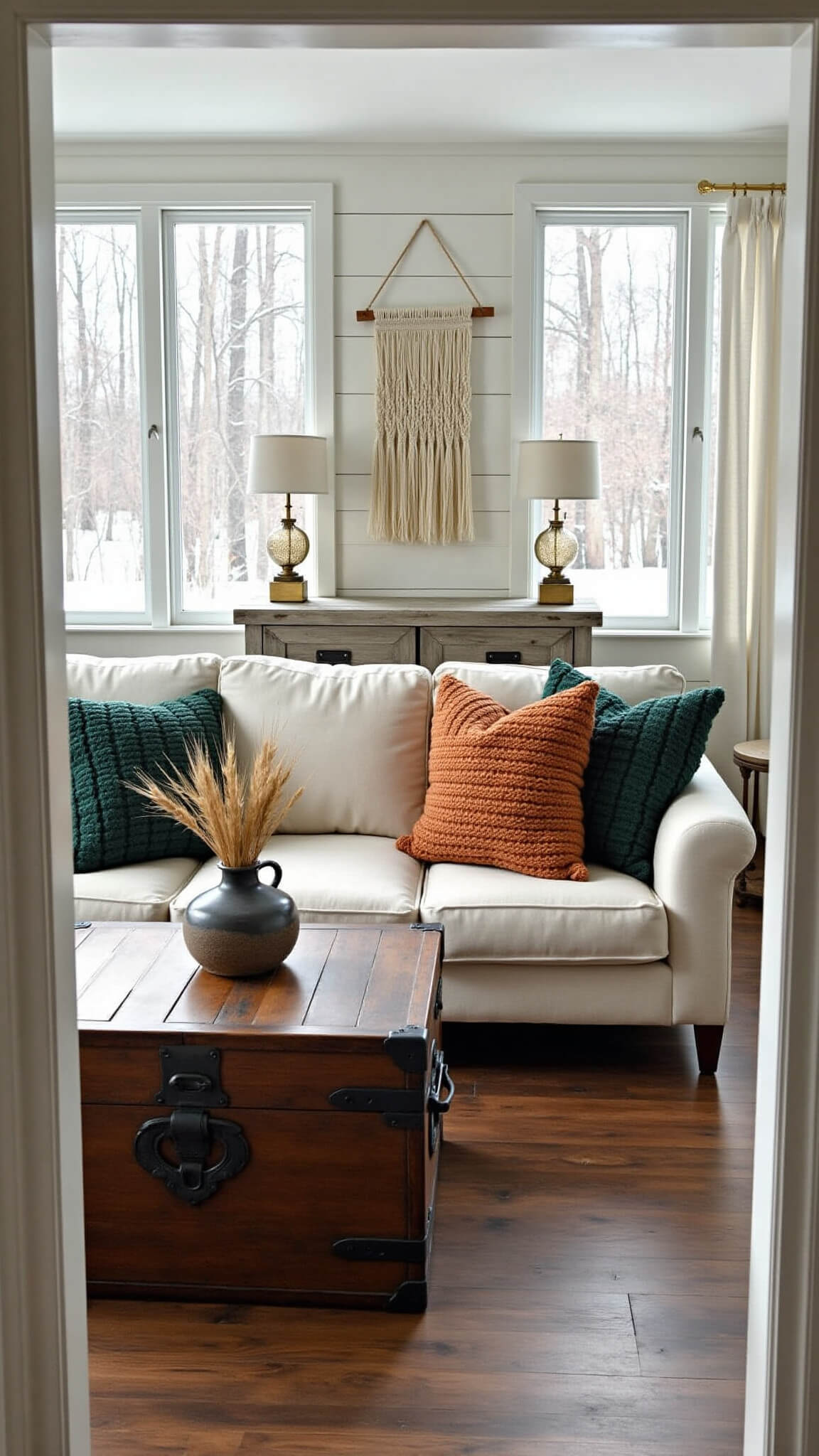 Cozy winter cabin interior with large windows showing snowy pines, oatmeal sectional, vintage trunk coffee table, and warm morning light.