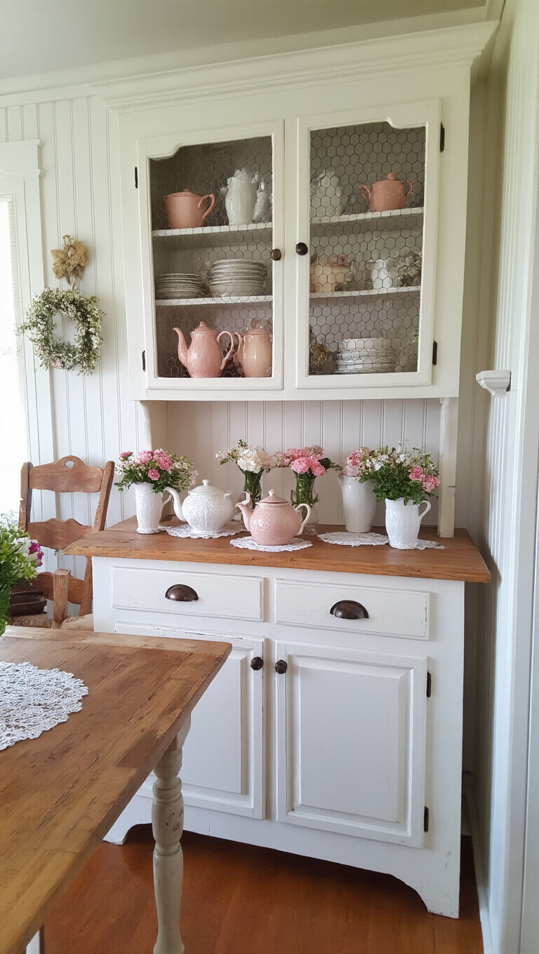 Cozy 8'x10' kitchenette with vintage teapots, built-in china cabinet with chicken wire fronts, white beadboard backsplash, worn wooden countertops, lace doilies, and fresh flowers in milk glass vases, softly lit during afternoon tea time.